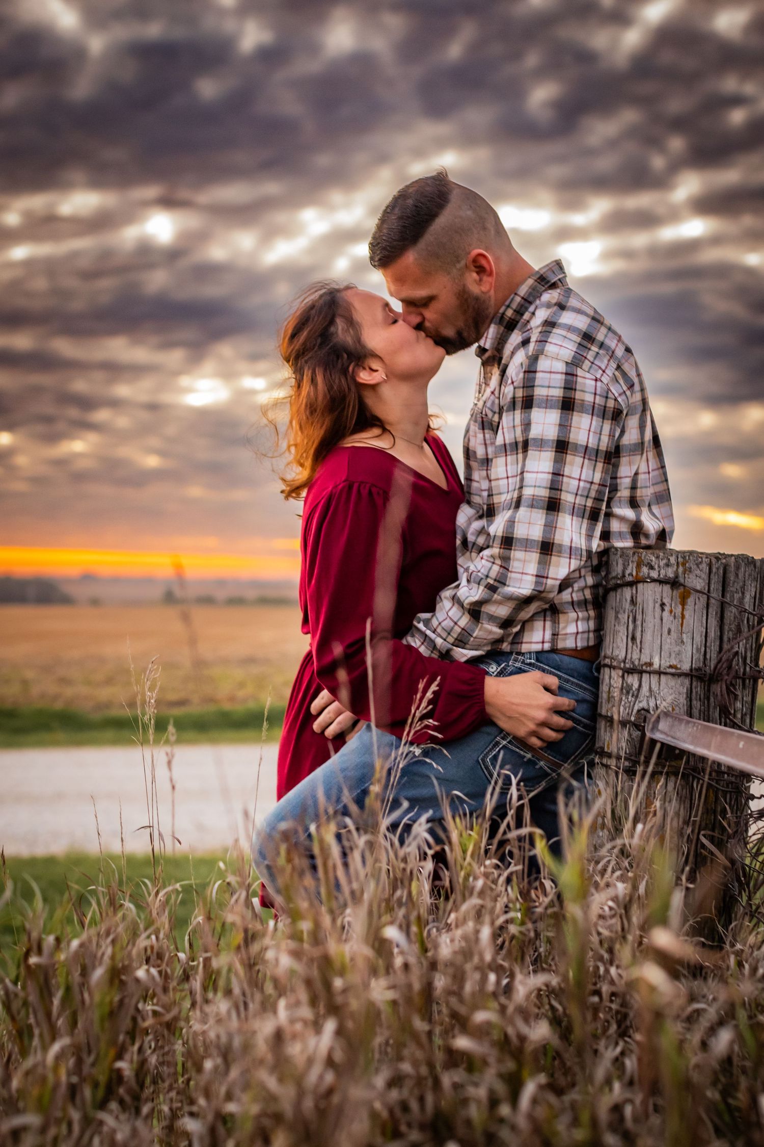 A romantic couple embraces in a field at sunset with dramatic cloudy skies.