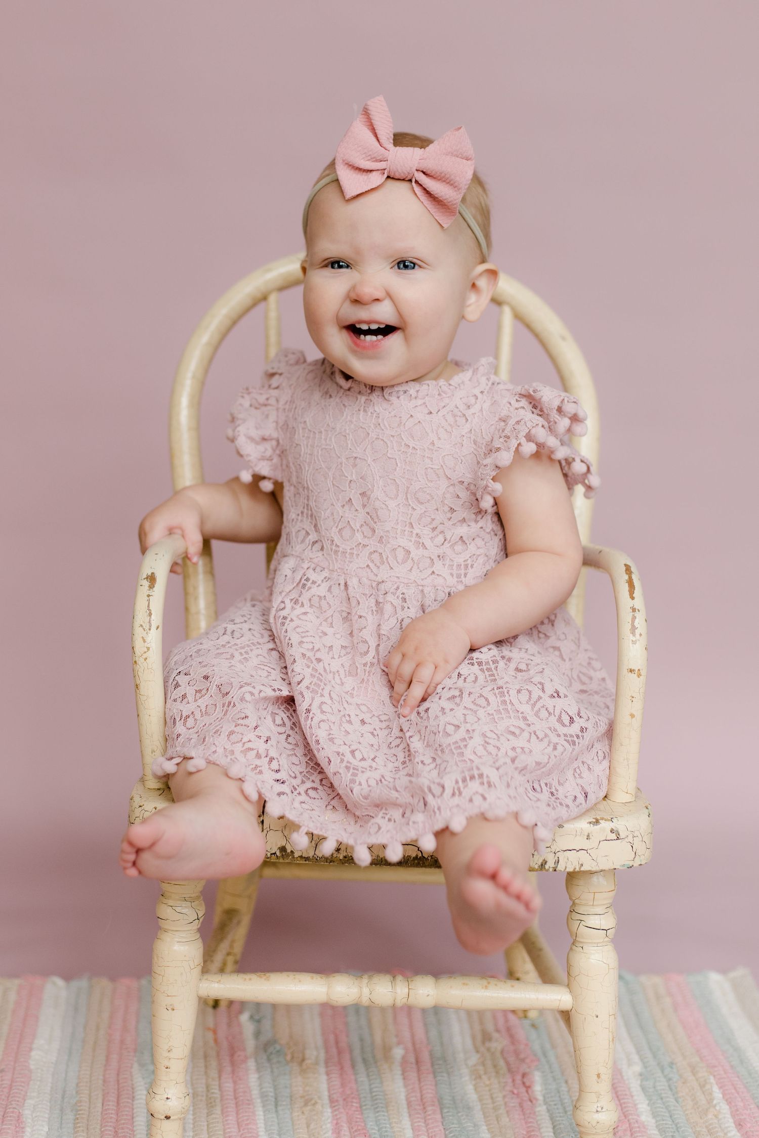A baby in a pink lace dress sitting on a vintage cream-colored chair.