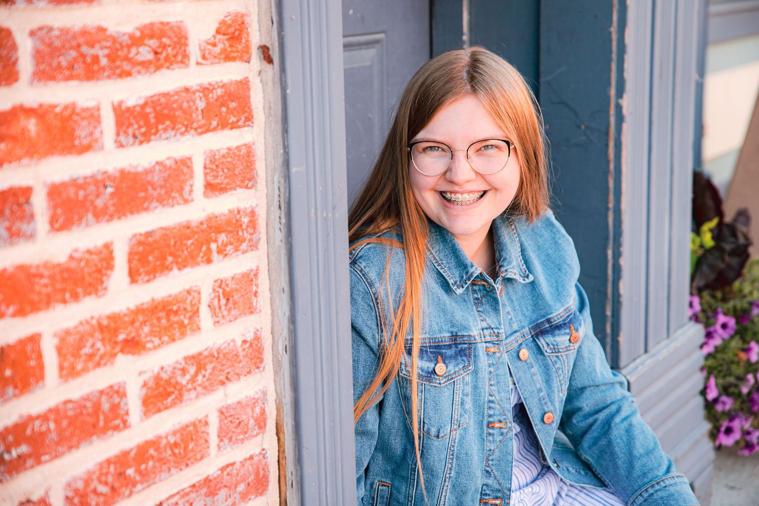 A person in glasses and denim jacket smiles while leaning against a brick wall with purple flowers nearby.
