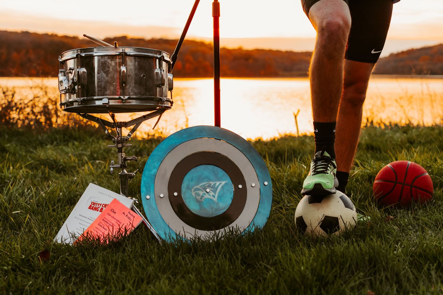 Sports equipment including a snare drum, archery target, soccer ball and cleats arranged on grass at sunset.