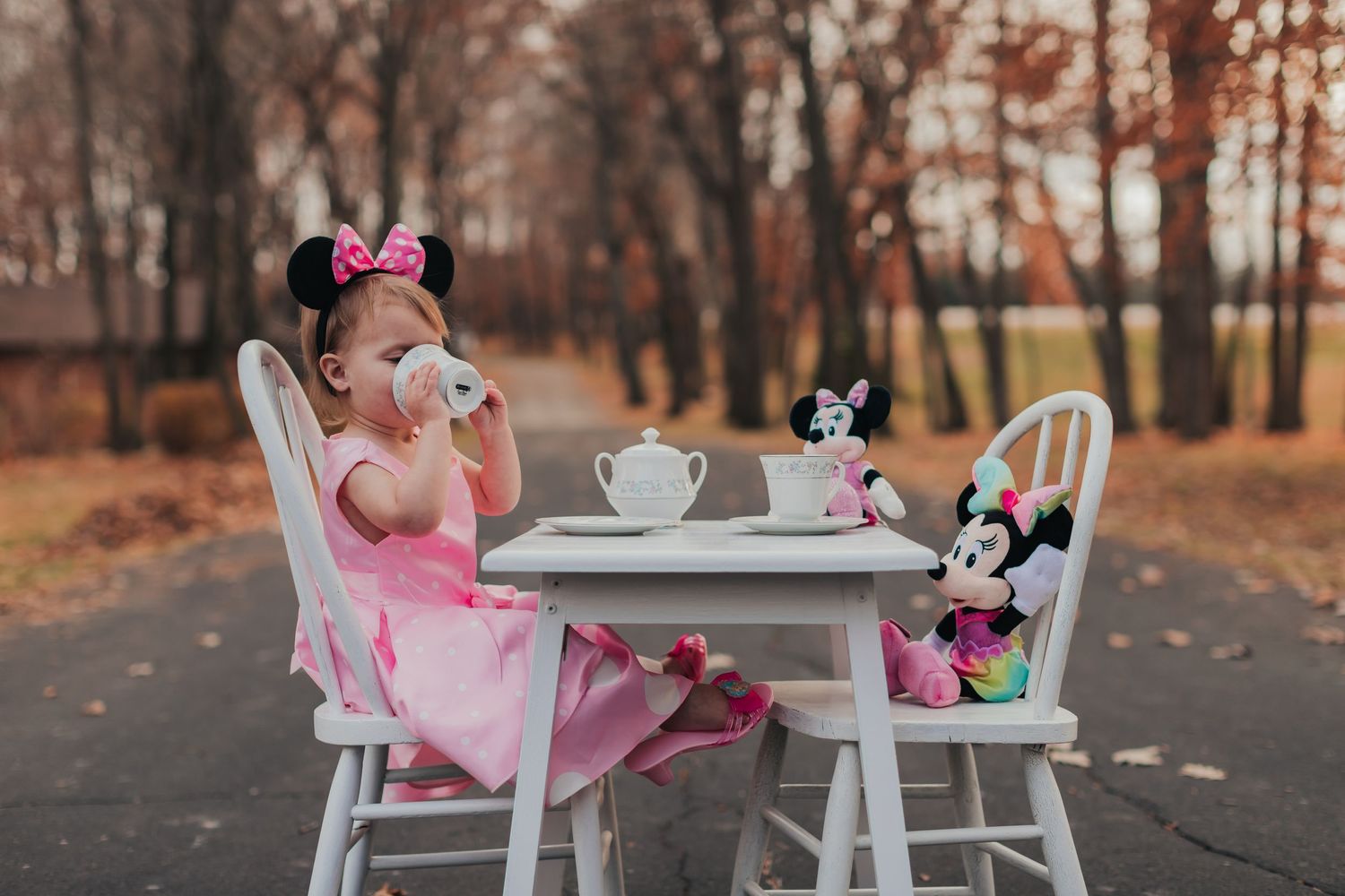 Child in pink dress having tea party with Minnie Mouse plush toys at white table.