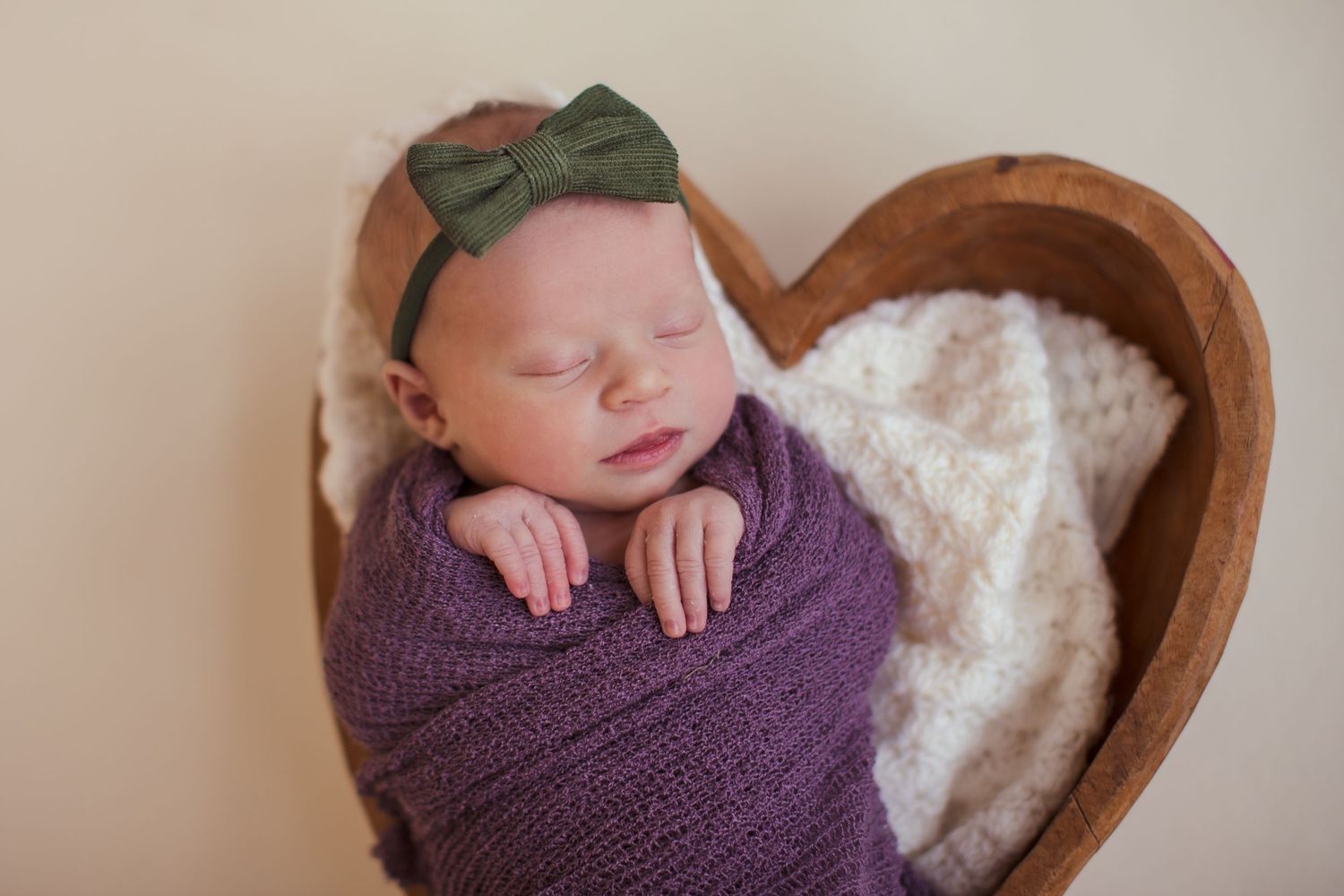 Sleeping newborn wrapped in purple swaddle with green bow in wooden heart-shaped basket.