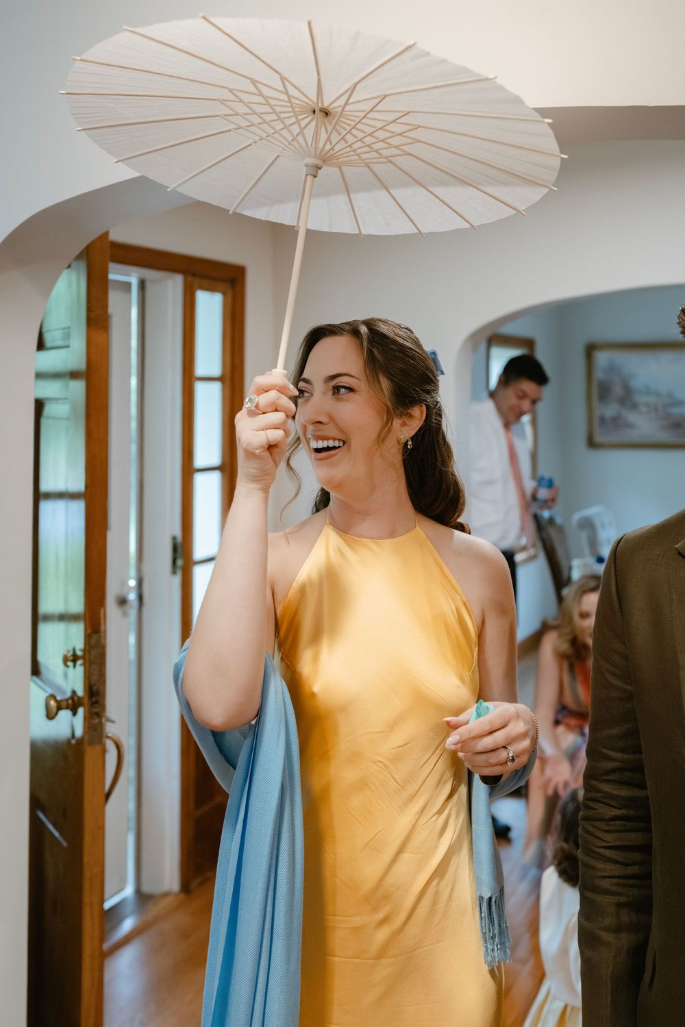 Someone in a yellow dress holds a white paper parasol while standing indoors near wooden-framed windows.