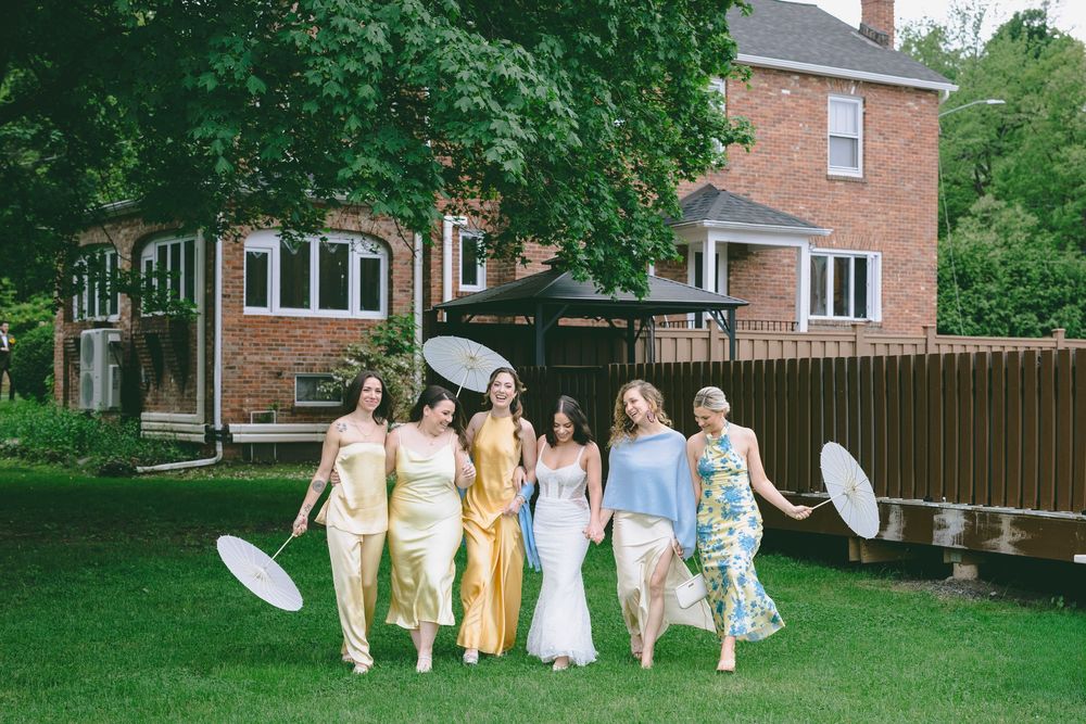 A group in coordinated pastel and yellow dresses walking together on a green lawn near brick buildings.