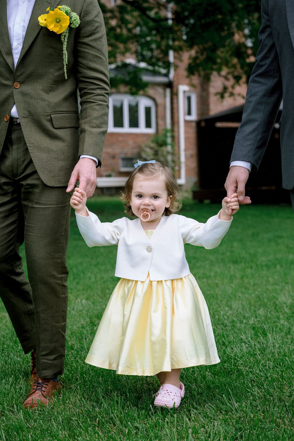 A young child in a white and yellow dress holds hands with adults in green suits on a grassy lawn.