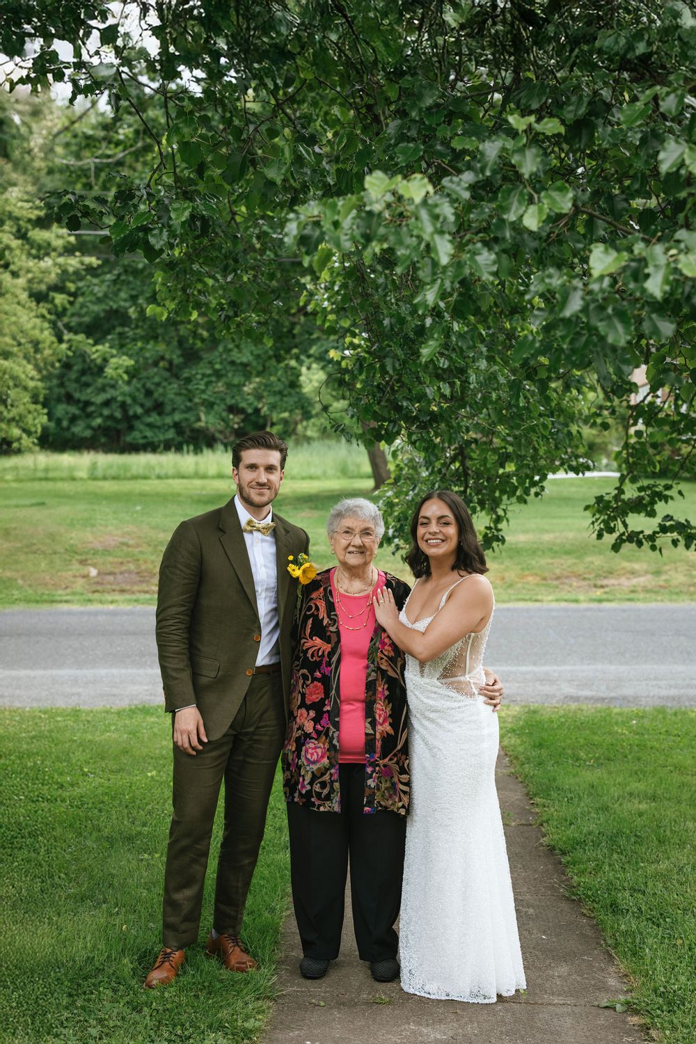 Three people pose together under a green tree canopy during an outdoor wedding celebration.
