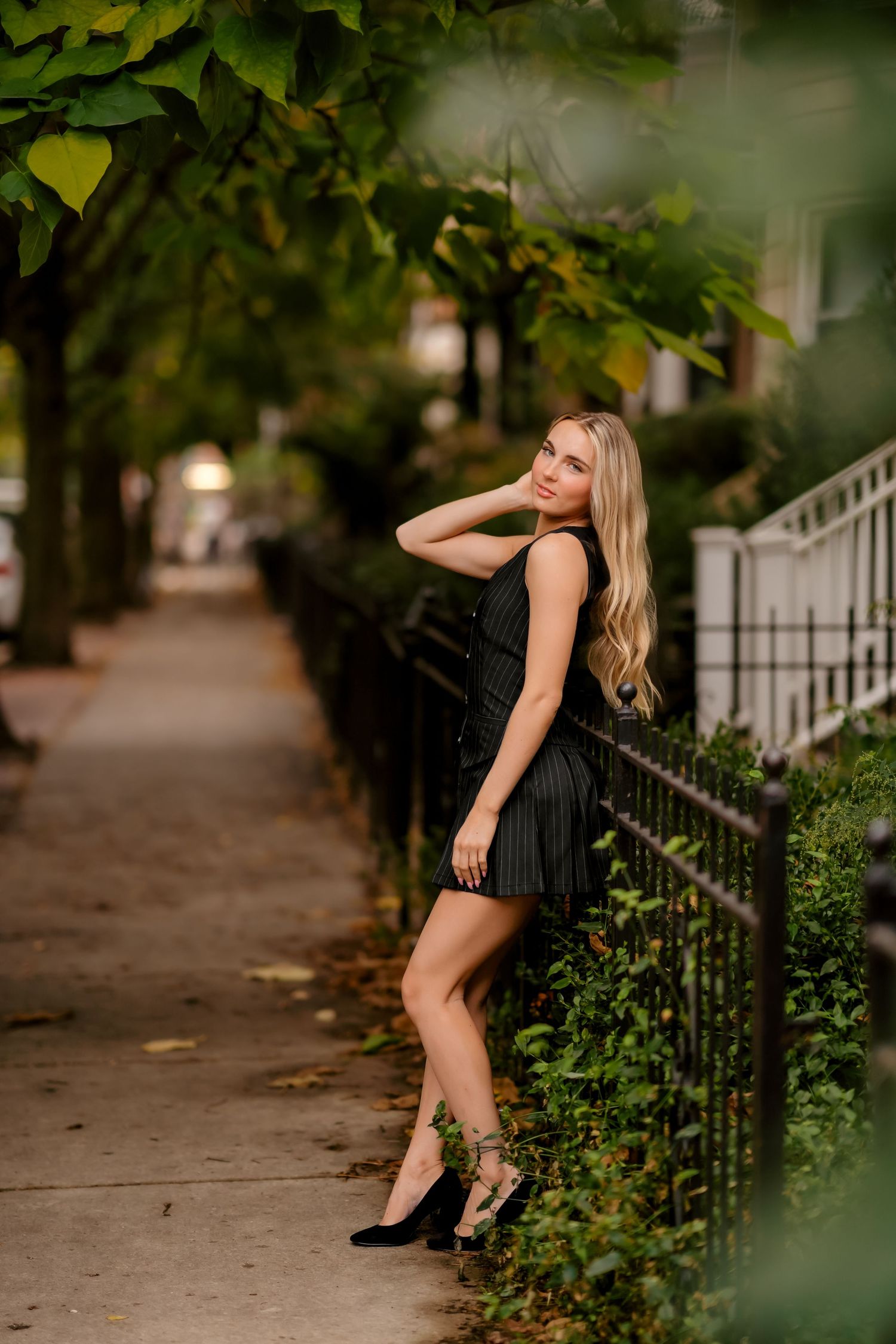 Fashion model in black dress poses elegantly against urban garden backdrop in evening photoshoot series.