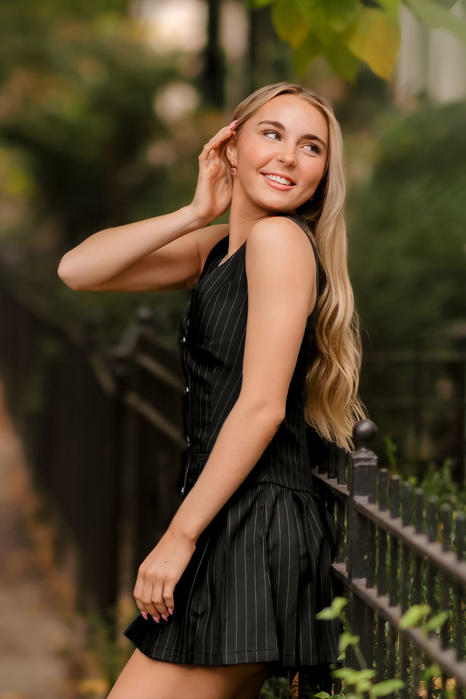 A woman in a black pinstripe top poses against an iron fence during a summer outdoor portrait session.
