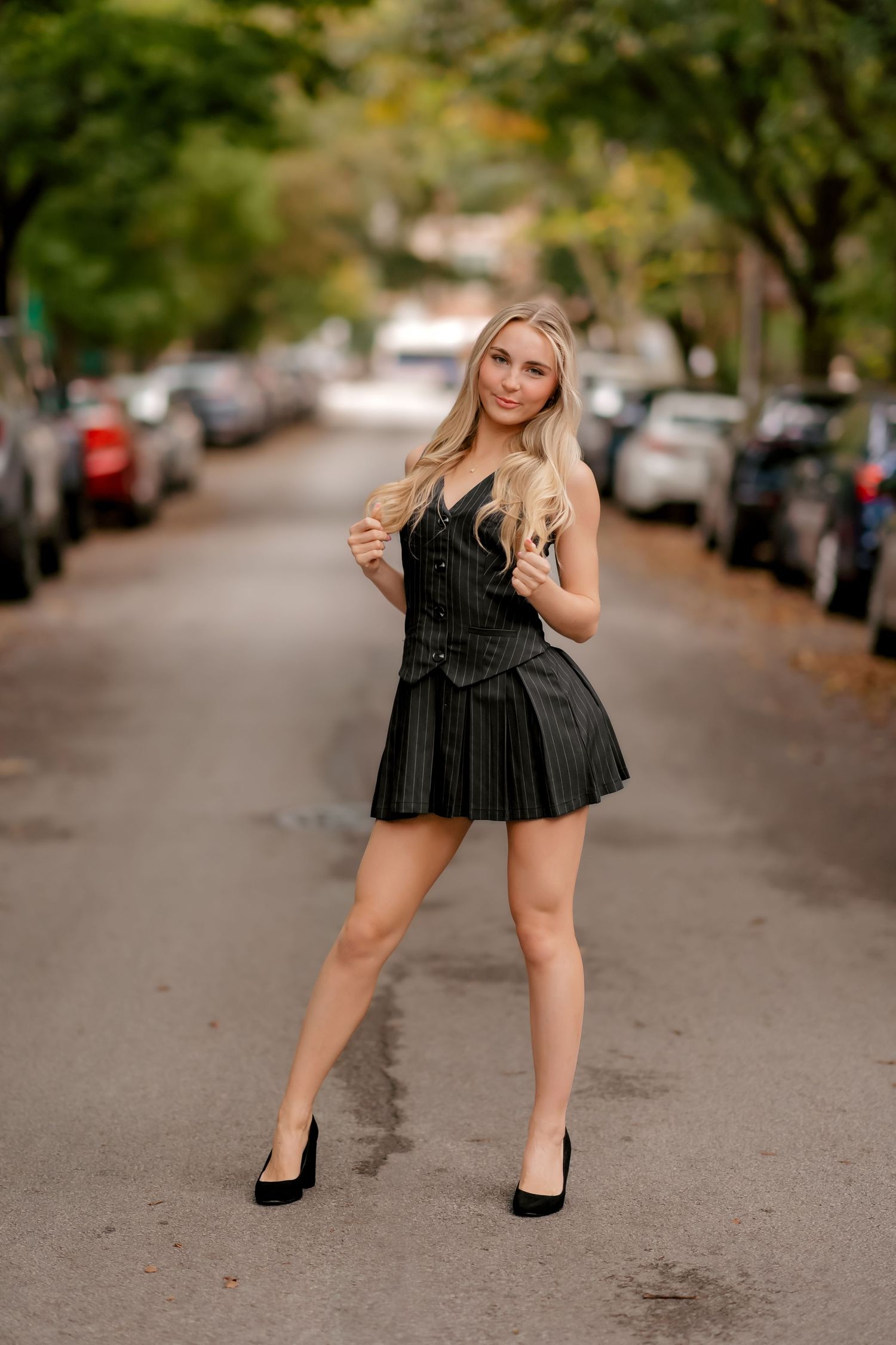 A woman in a black dress poses on a tree-lined street with parked cars in the background.