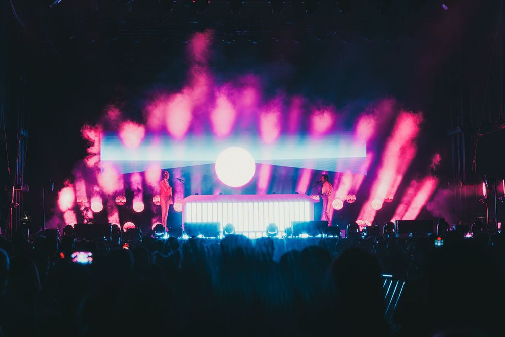 Music festival panorama showing pink and purple stage lights glowing across a dark outdoor venue.
