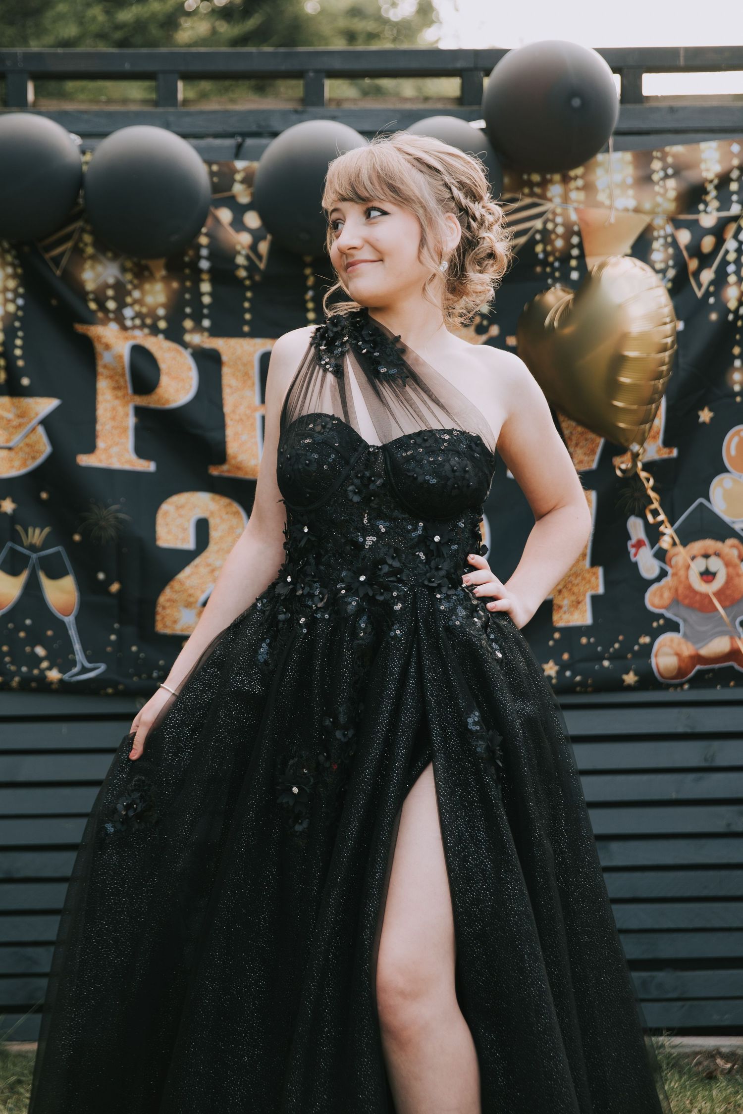 A person in a sparkly black formal gown poses against a dark wall with decorative lettering.