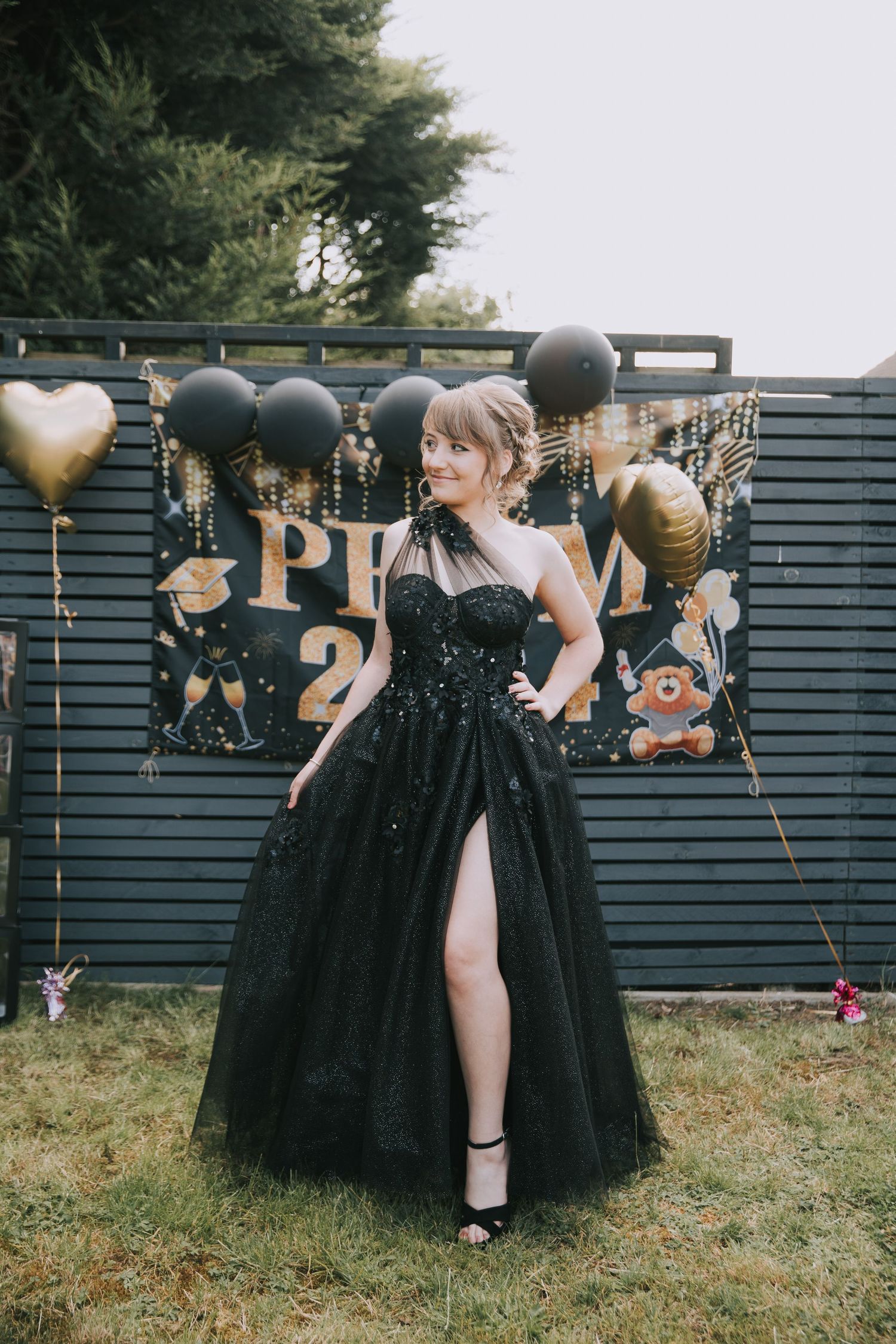 A group in elegant black dresses poses with gold balloons against a dark fence at an outdoor celebration.