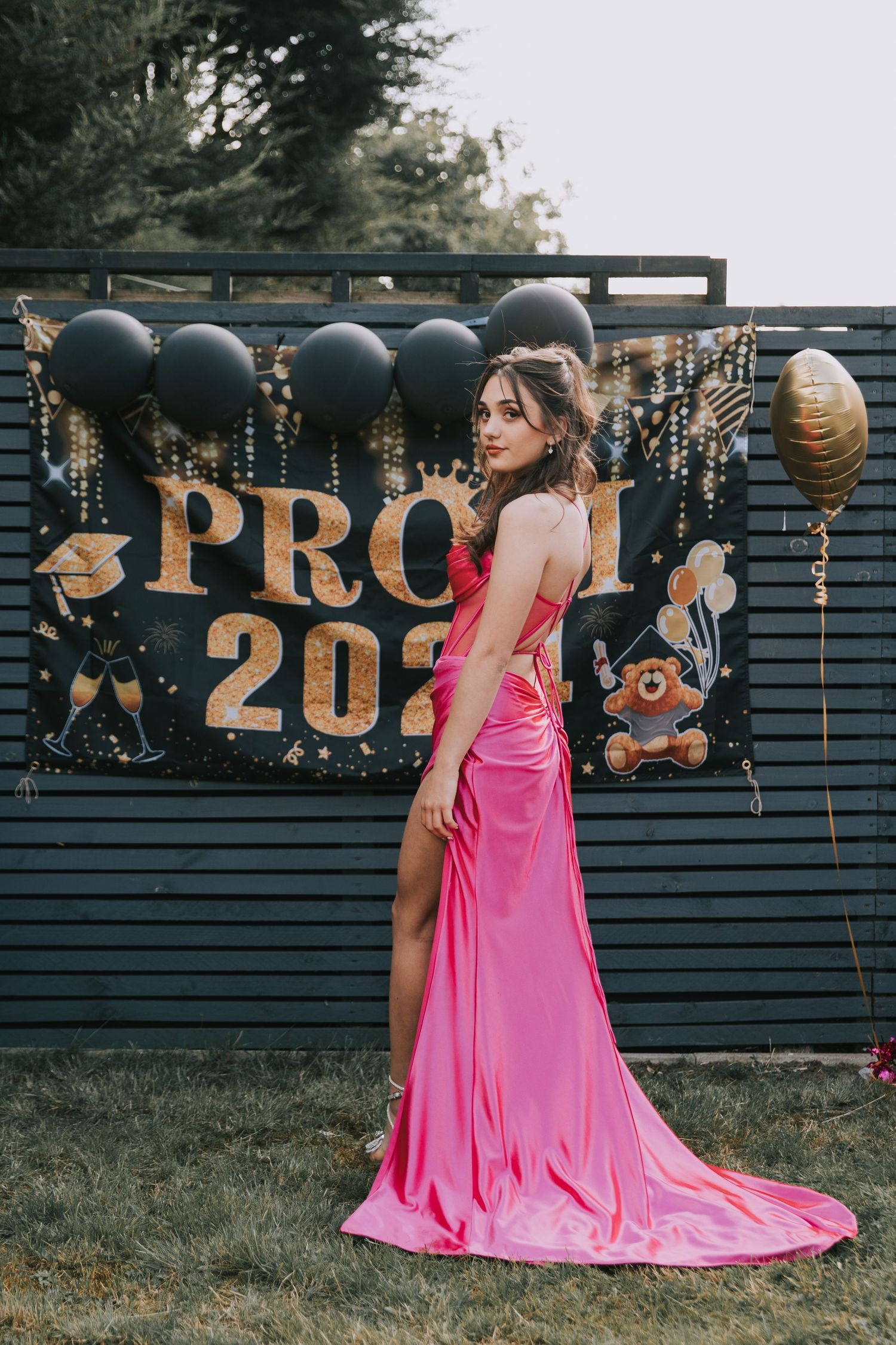 A figure in a flowing hot pink dress poses against a dark fence decorated with gold numbers and hanging lanterns.