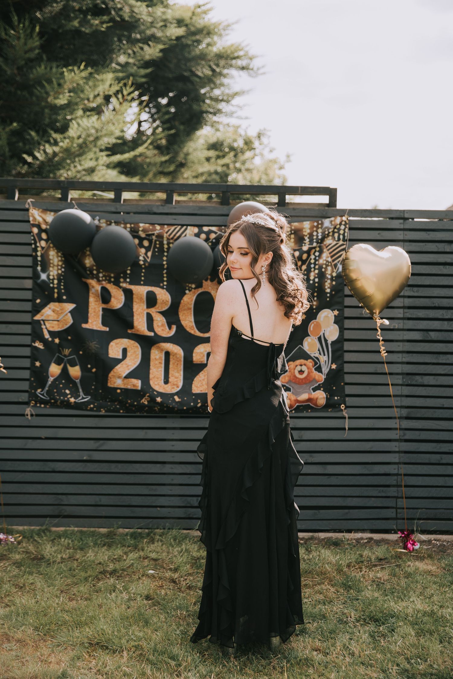 Elegant black prom dress photographed against dark green fence with gold 'PROM 2021' banner and festive balloons.