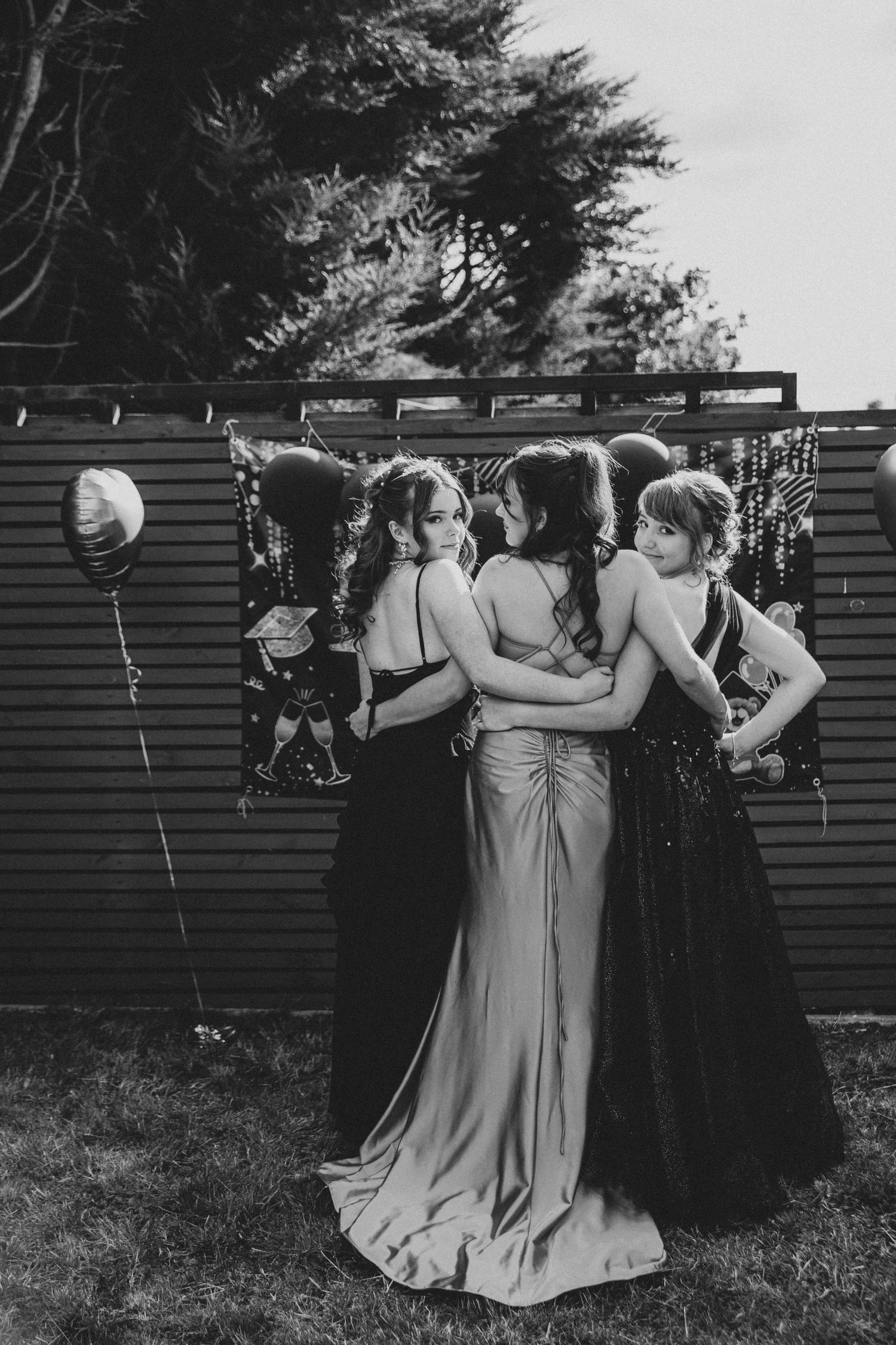A group of bridesmaids in matching dresses walk arm in arm through a garden in black and white.