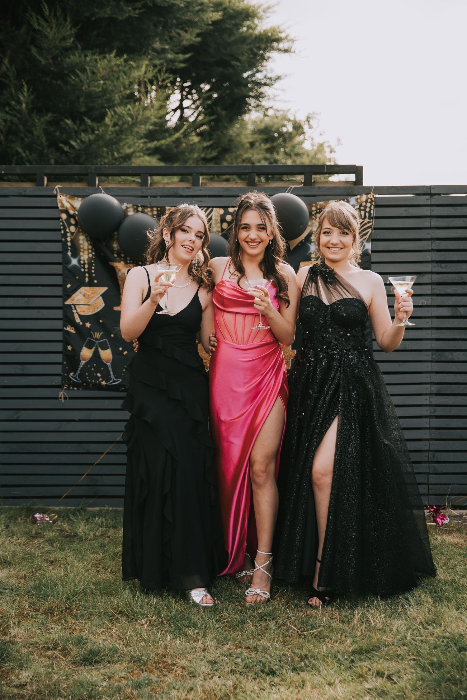 A group in formal attire poses playfully outdoors, with some wearing black dresses and one in pink.