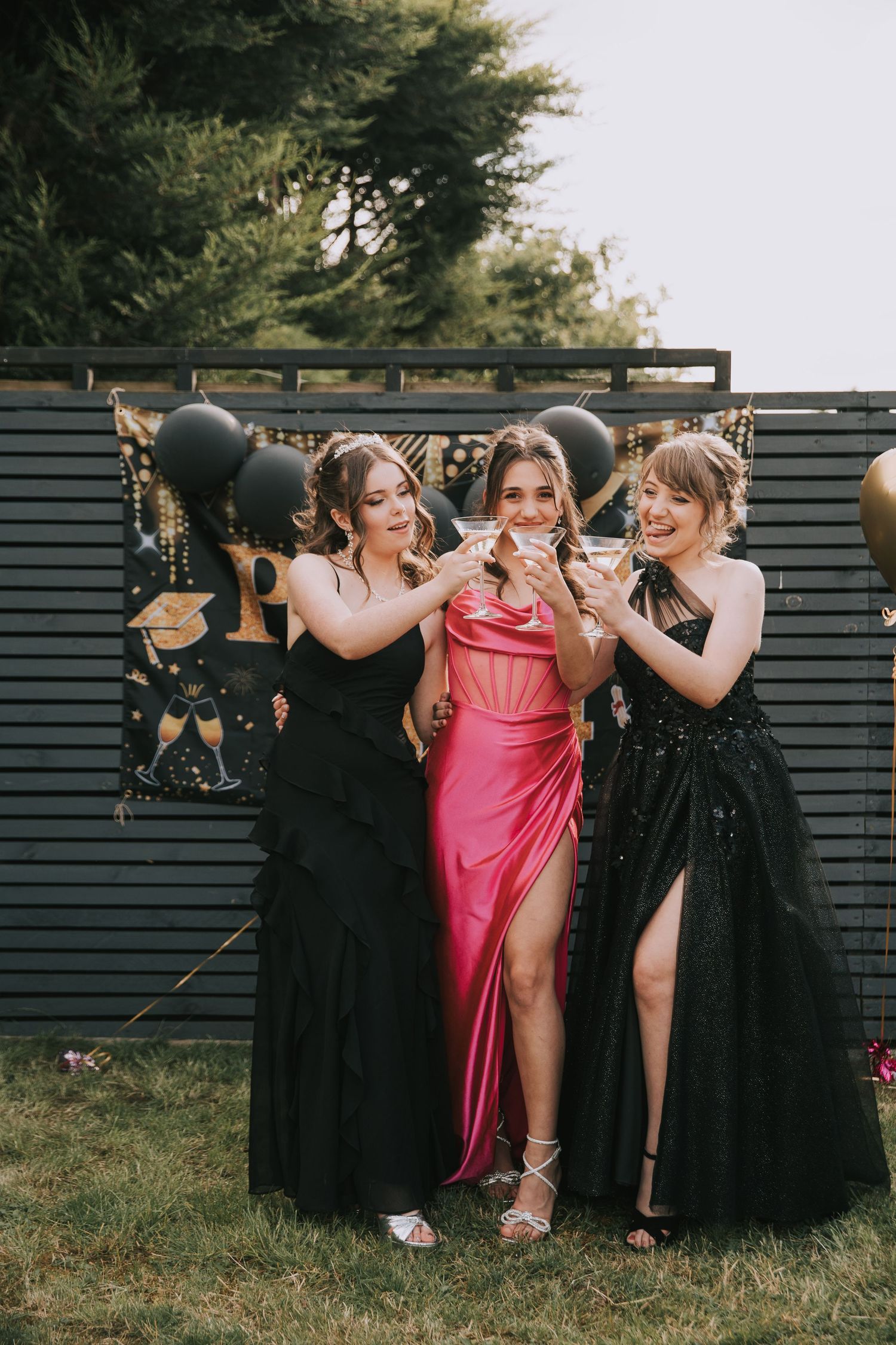 A group in formal attire poses playfully with black umbrellas outdoors against a dark fence.