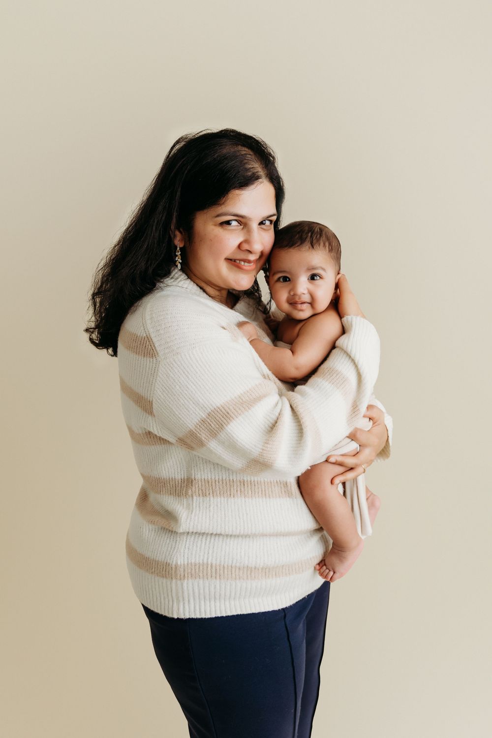 A parent in a cream striped sweater holds a smiling baby wearing white against a neutral background.