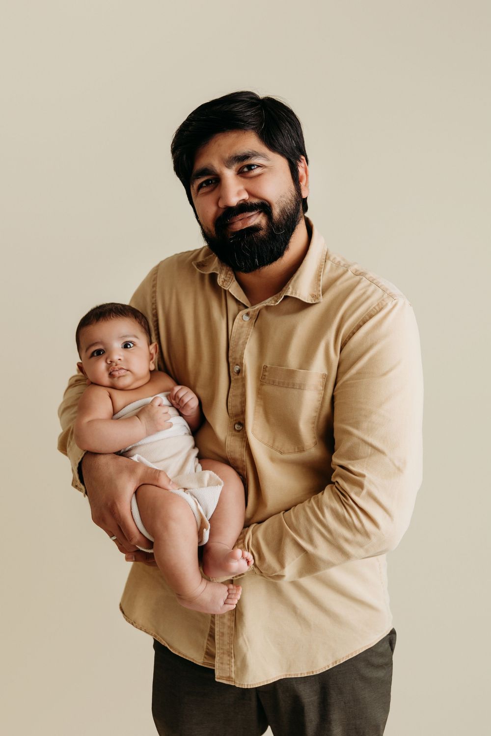 A parent in a beige shirt holds a small baby in white clothing against a neutral studio backdrop.