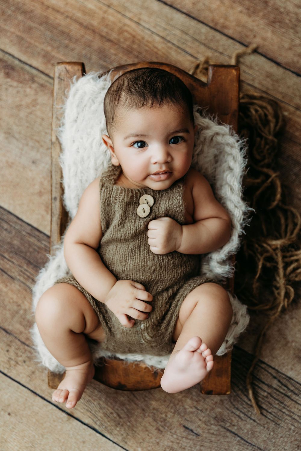 Baby wearing knit romper lies on wooden floor with white fluffy blanket in professional photography setup.