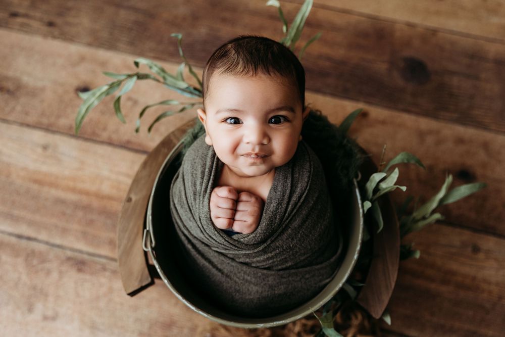 A newborn baby wrapped in a dark gray blanket poses in a wooden bowl with green foliage accents for a professional photo.