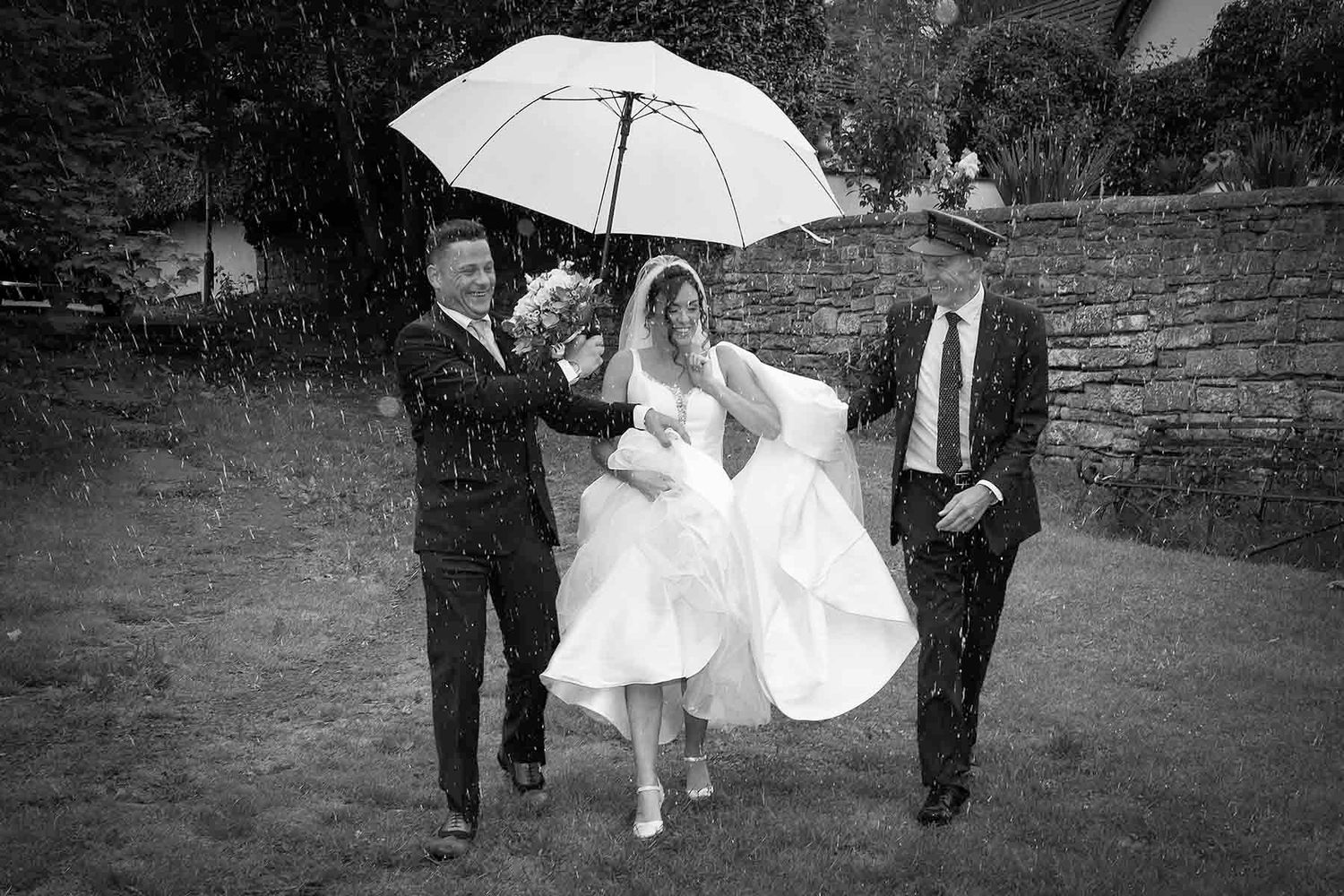 A wedding party walks through a garden while holding white umbrellas on an overcast day.