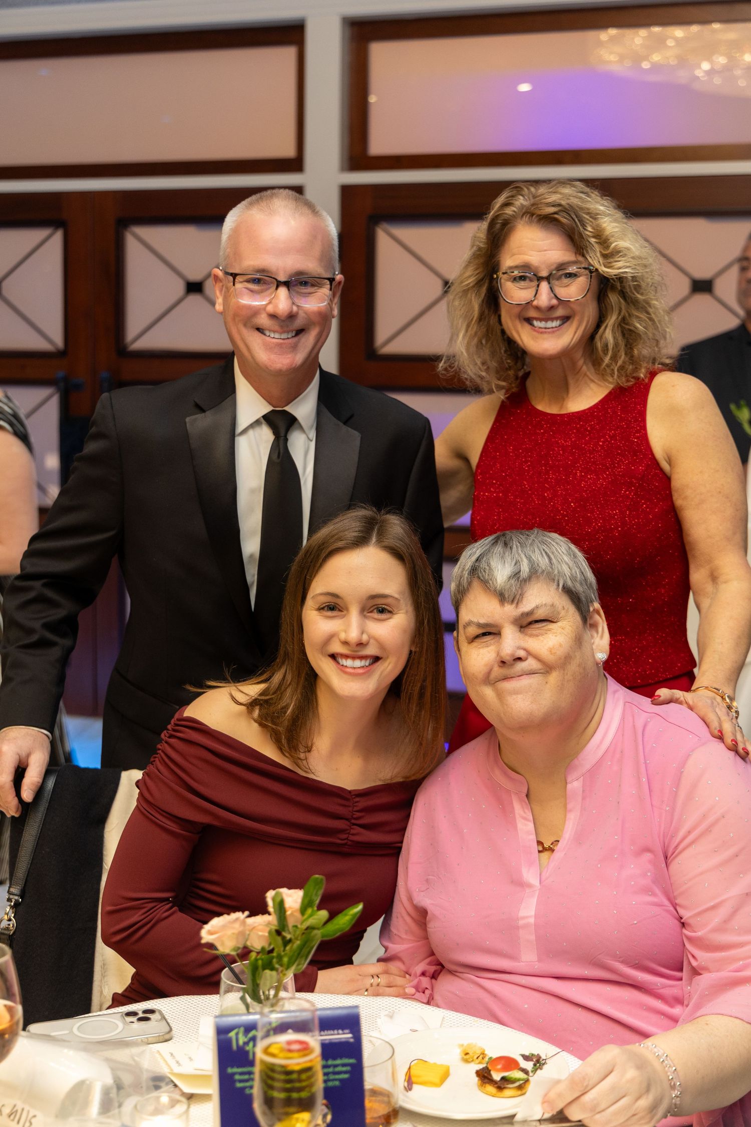 A group poses together at a formal dinner event, with two people standing and two seated at a table with place settings.