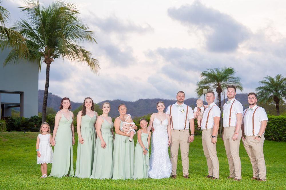 Wedding party in mint green and white attire poses outdoors with palm trees and mountains in background.