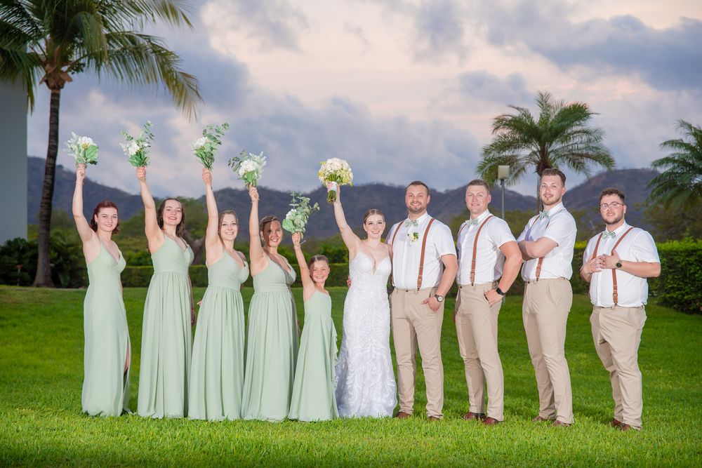 Wedding party poses together on a lush green lawn with palm trees and tropical resort buildings in the background.