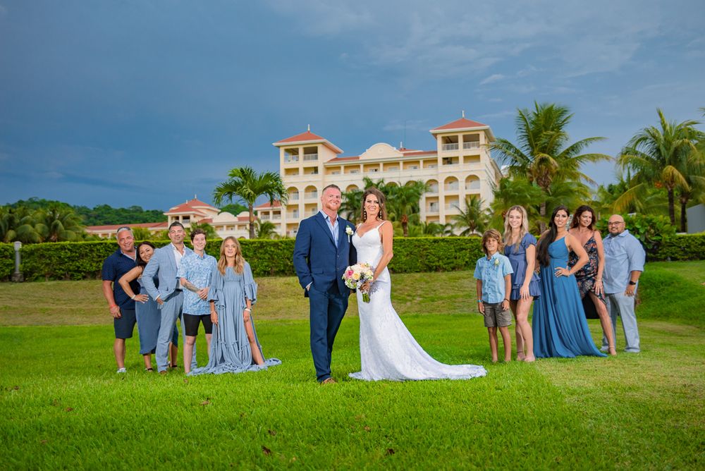 Wedding party in formal attire poses together on a lush green golf course with palm trees and resort buildings in background.
