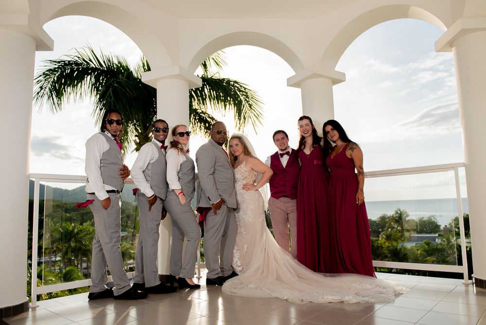 Wedding party stands in an elegant white columned pavilion overlooking a tropical beach landscape during ceremony.