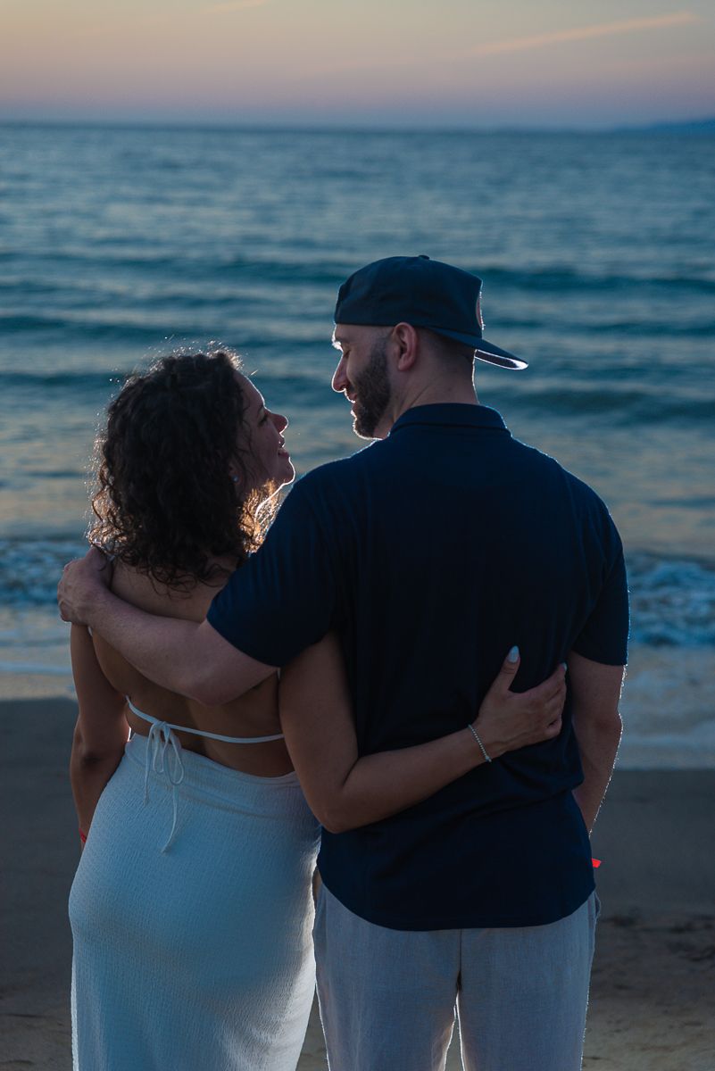 A romantic couple embraces while watching the sunset over the ocean on a peaceful beach.