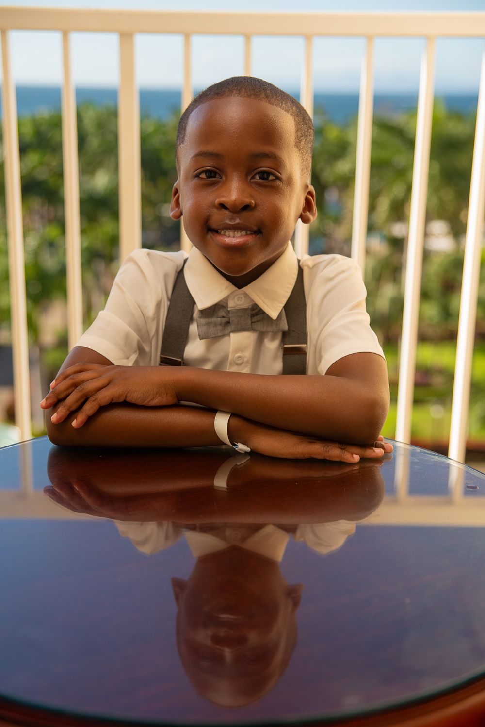 Young student in formal attire with bow tie and suspenders smiles while leaning on reflective table with ocean view behind.