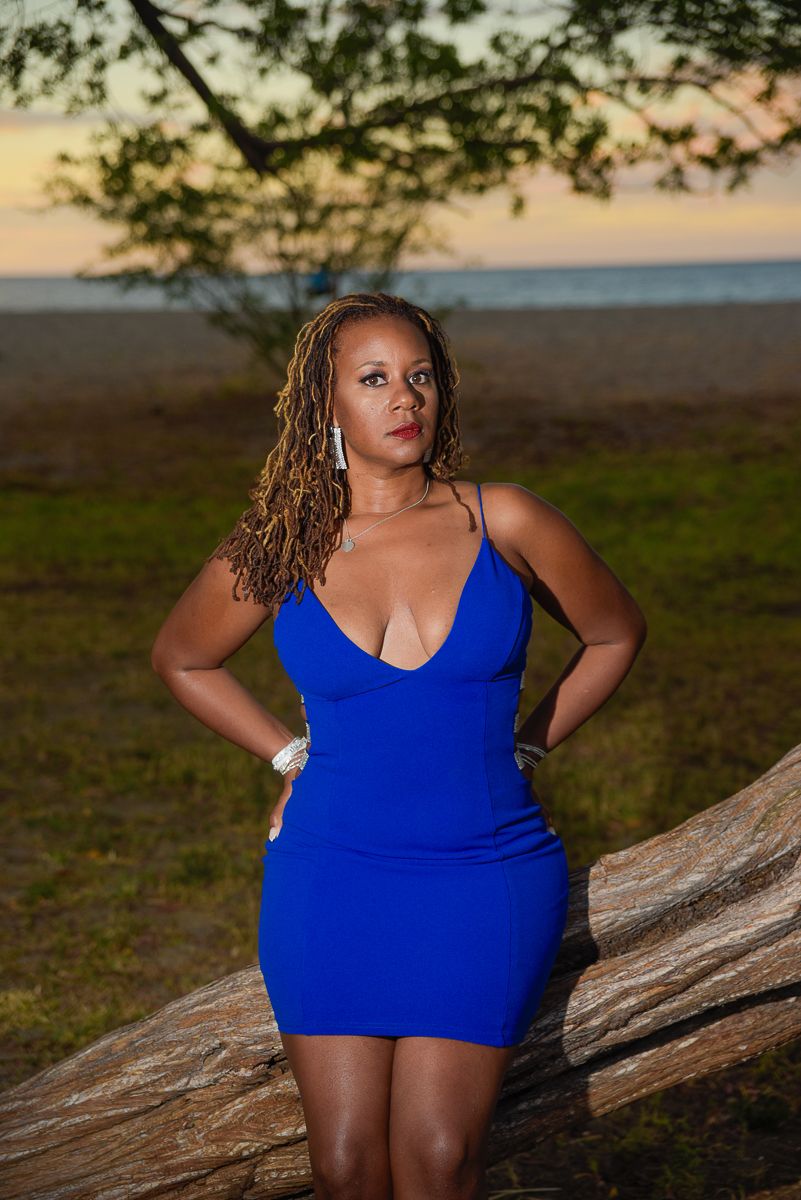 A person in a royal blue bodycon dress poses against a fallen tree trunk at sunset by the beach.