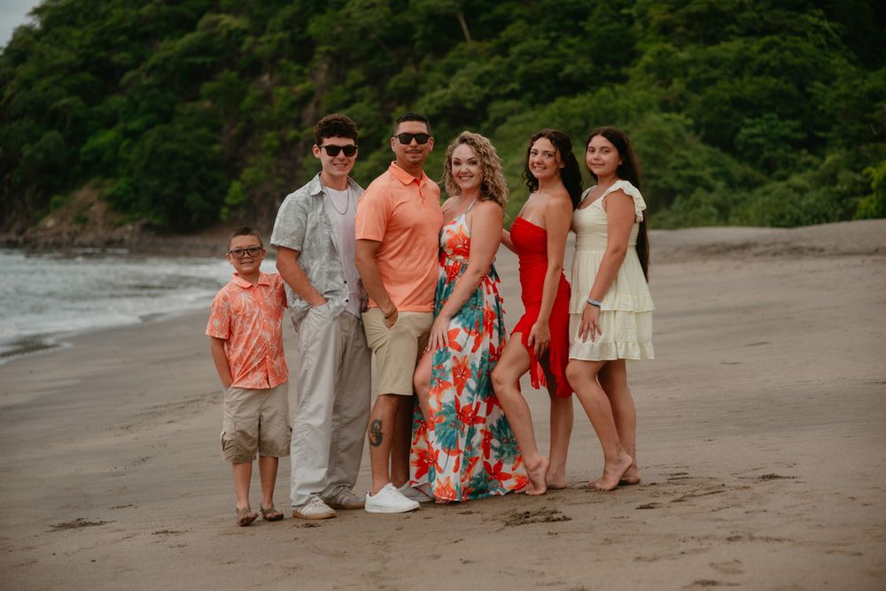 A group in coordinated coral and beige outfits poses together on a scenic beach with lush greenery in the background.