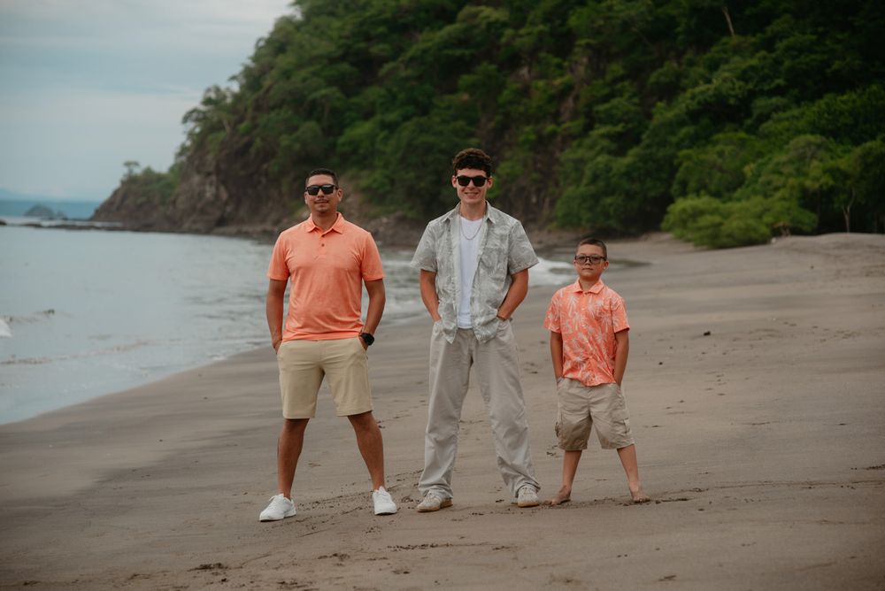 A group walks along a scenic tropical beach with lush green cliffs and ocean views in the background.