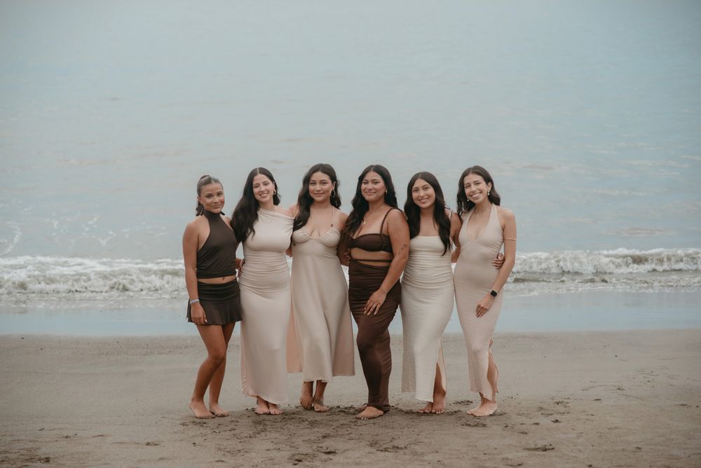 A group poses casually together on a sandy beach with gentle waves and cloudy skies in the background.