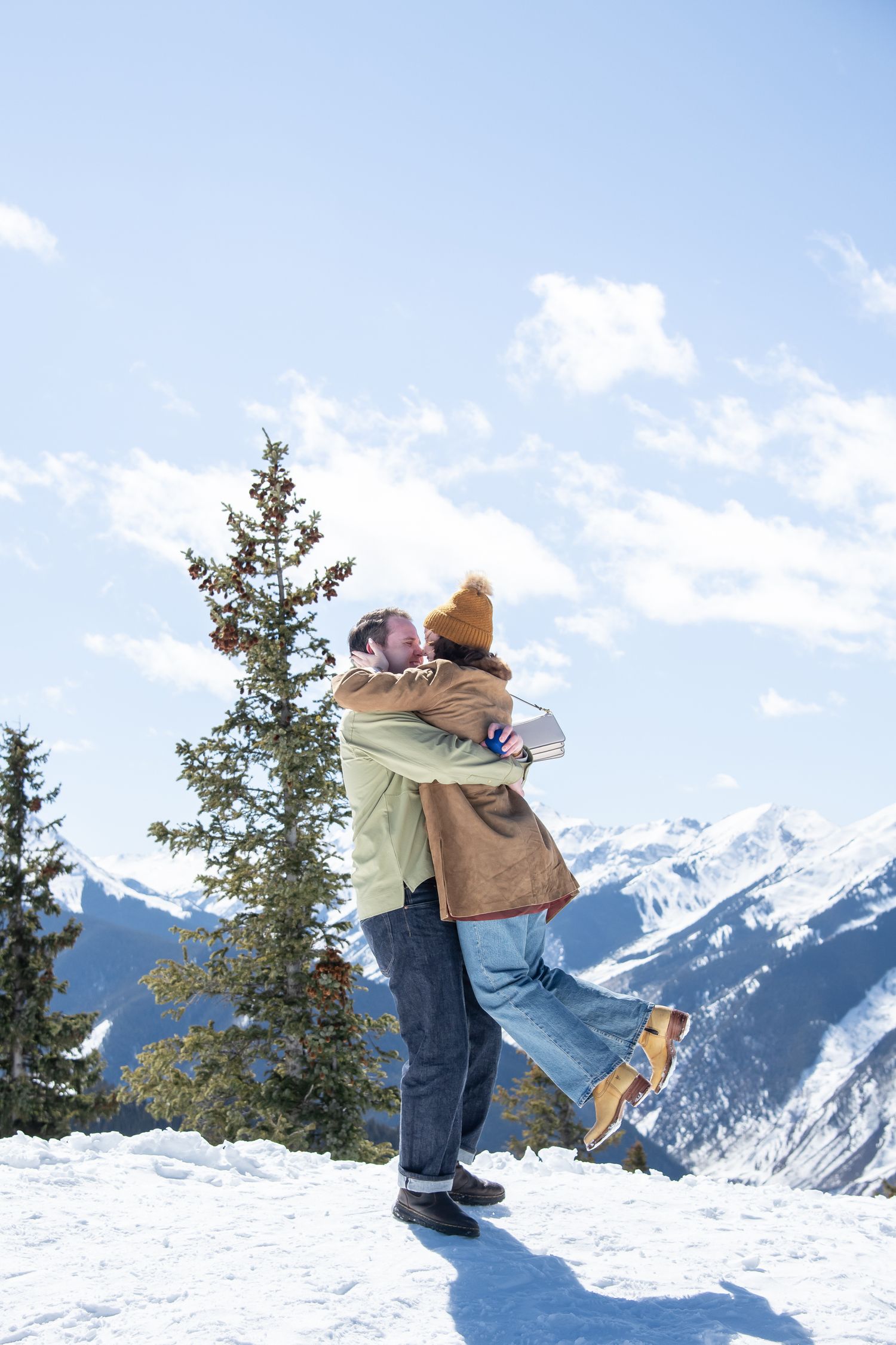 Couple embracing on snowy mountain peak with evergreen trees and snow-capped peaks in the background for a winter proposal in Aspen CO.