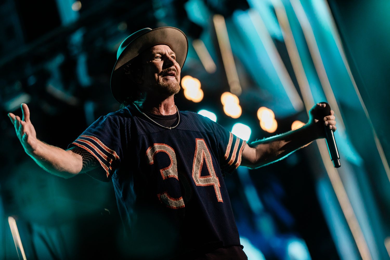 A performer in a number 24 jersey sings on stage with arms outstretched under dramatic turquoise lighting.