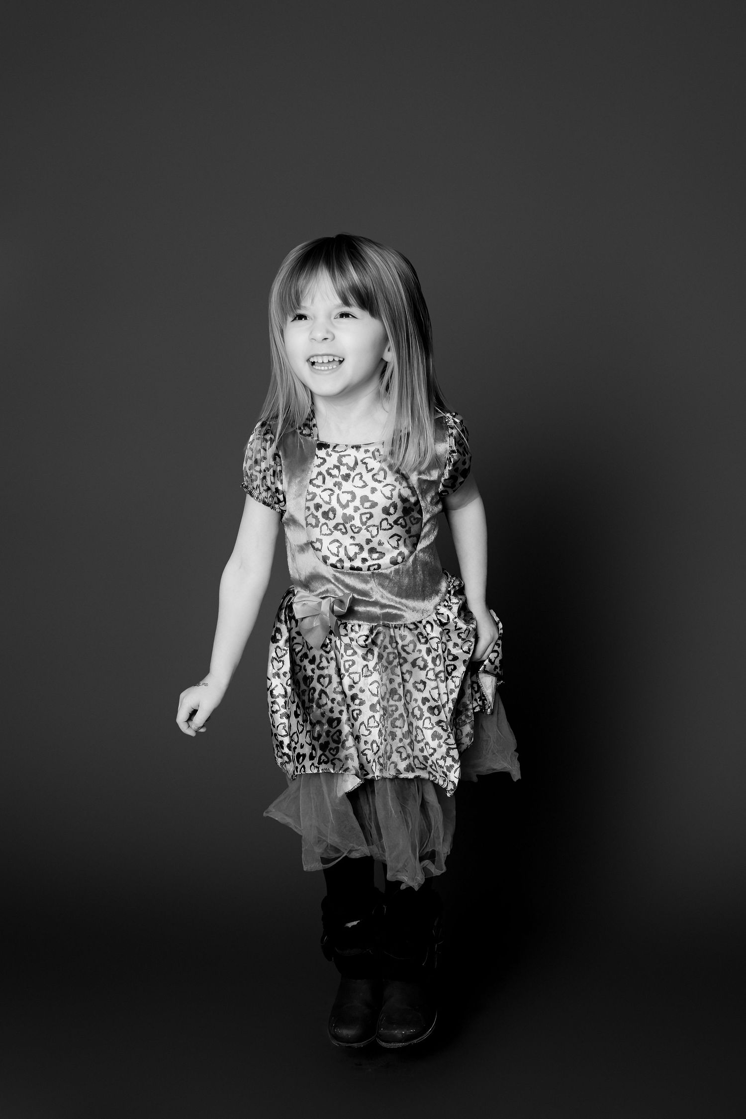 A child wearing a leopard print top and dark pants poses playfully against a dark background in a black and white photo series.
