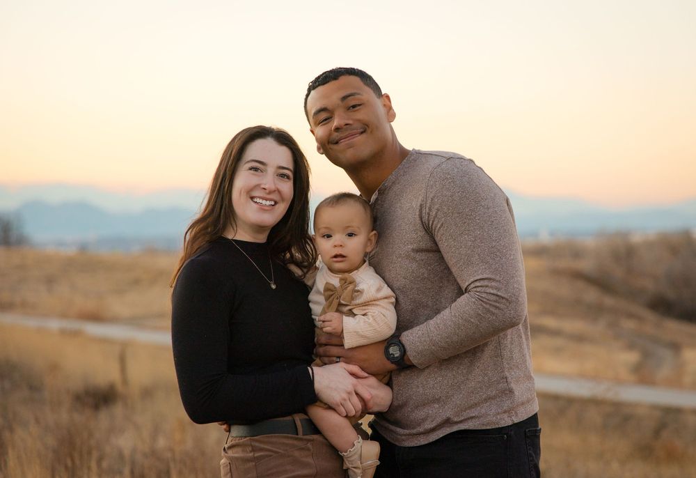 Family of three posing together in casual attire during a warm sunset photoshoot in an outdoor desert setting.