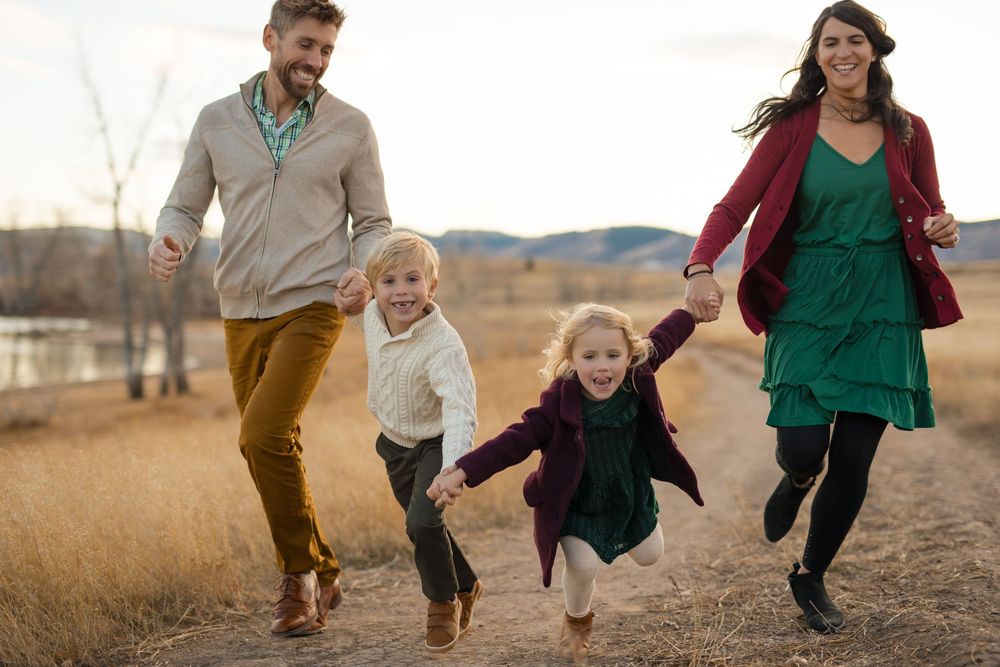 A family joyfully runs and holds hands together on a dirt path through a golden field with mountains in the background.
