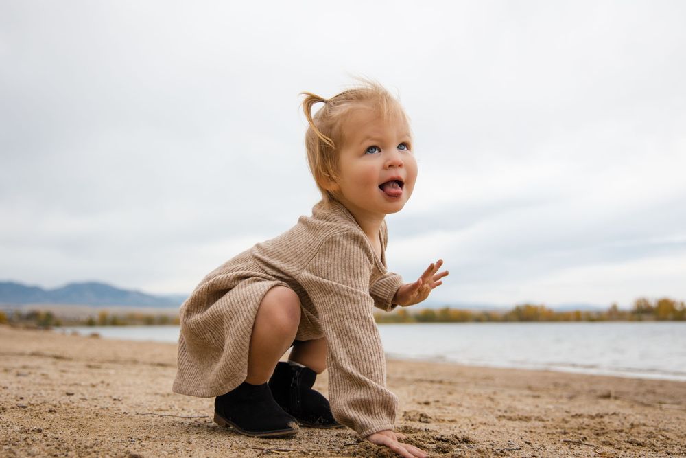 A toddler in a beige sweater crawls and plays on a sandy beach near mountains and water on a cloudy day.