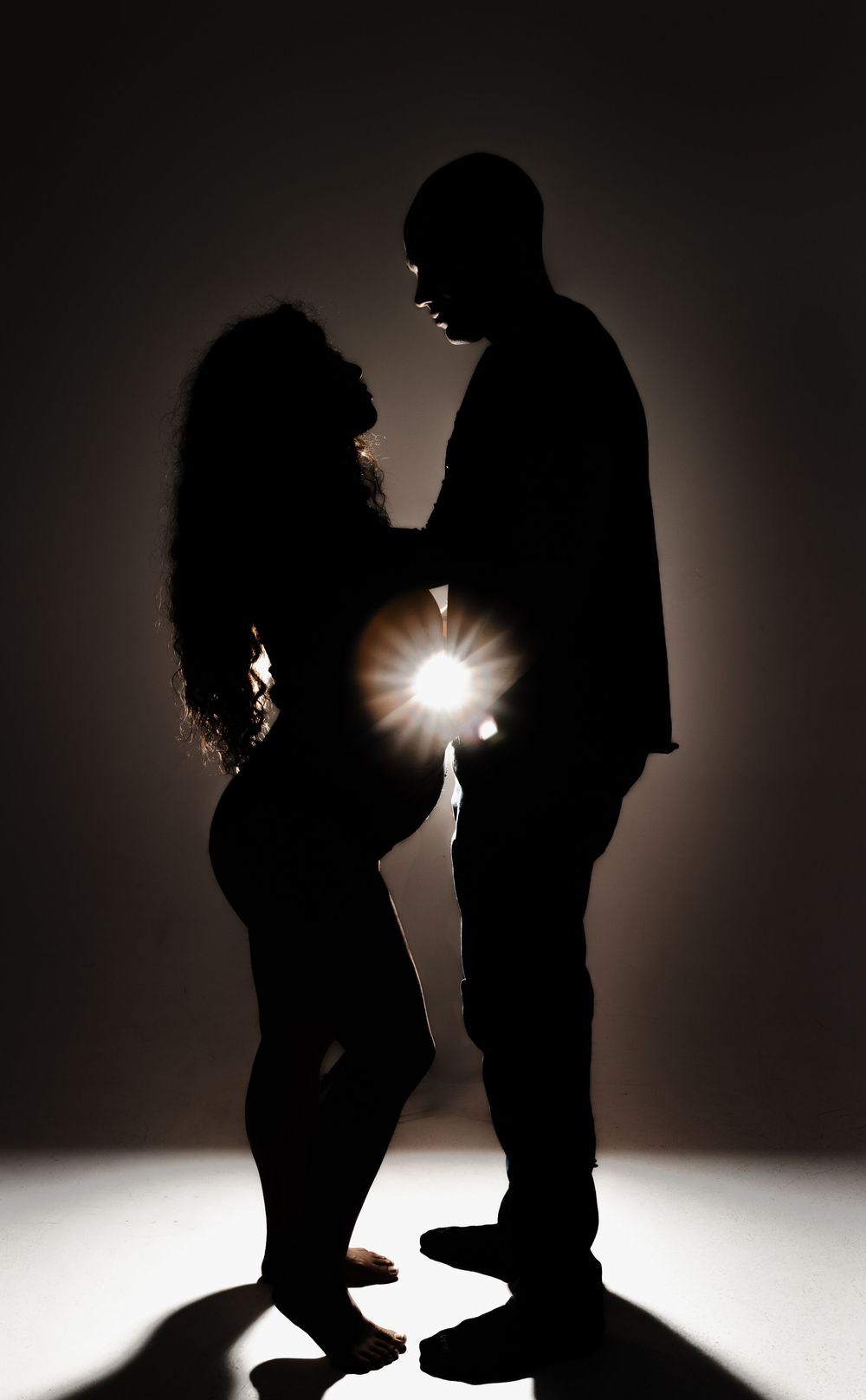 Silhouetted couple stands facing each other with dramatic backlight creating a romantic atmosphere in dark studio setting.