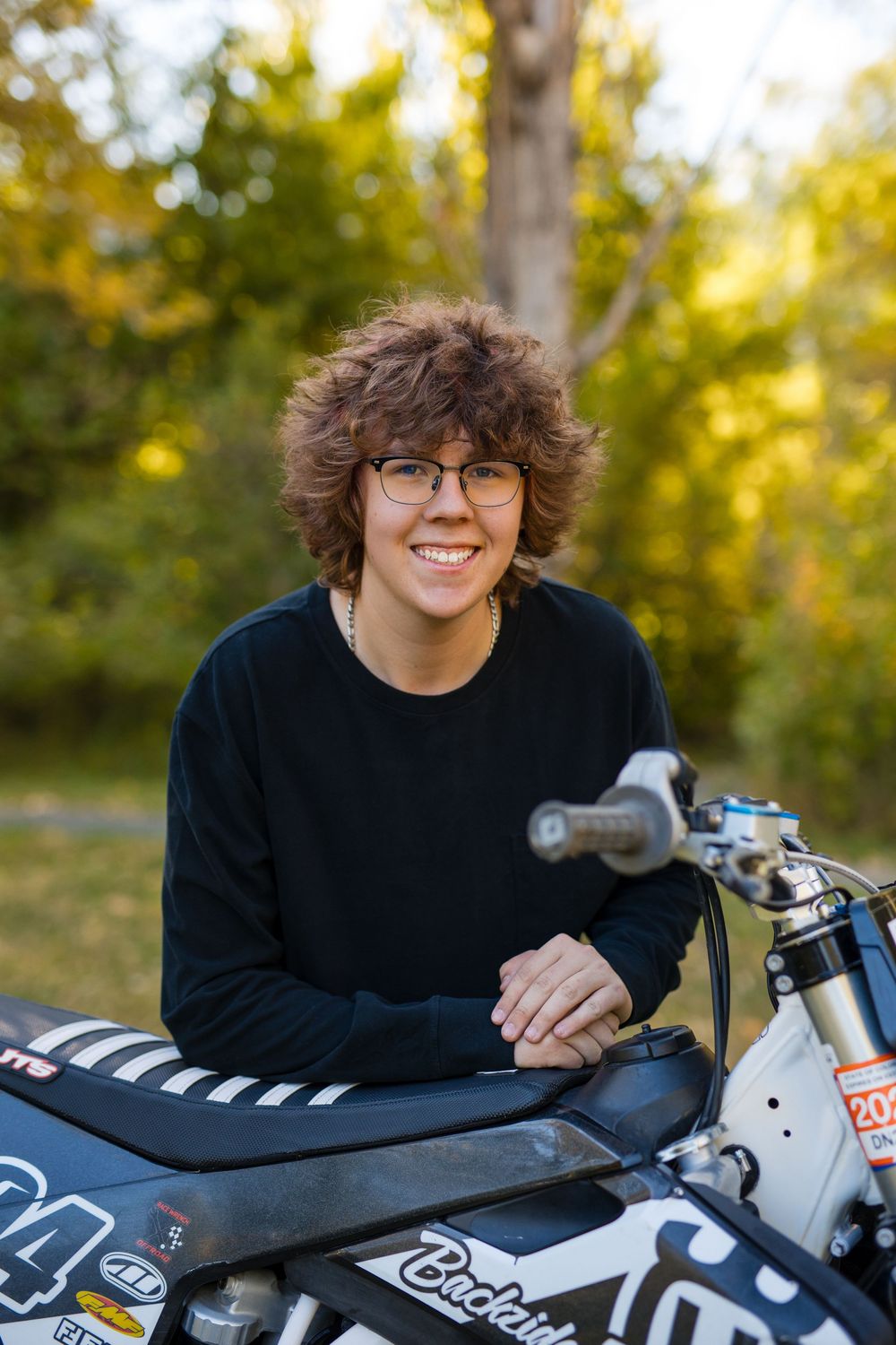 Person in black sweater and glasses poses on a motorcycle outdoors with autumn foliage in background.