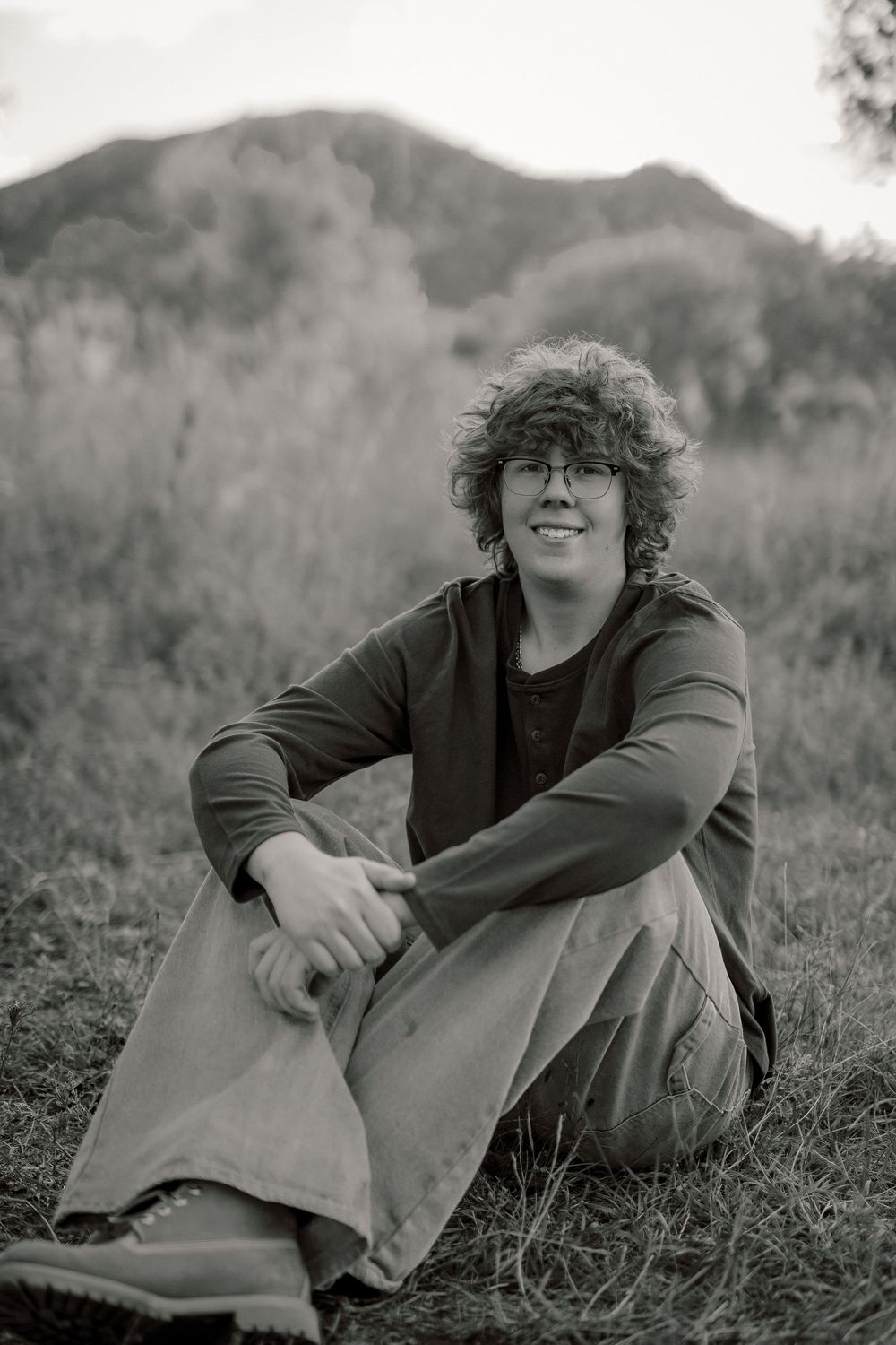 Black and white portrait of someone with curly hair sitting in grass wearing a dark sweater and smiling warmly.