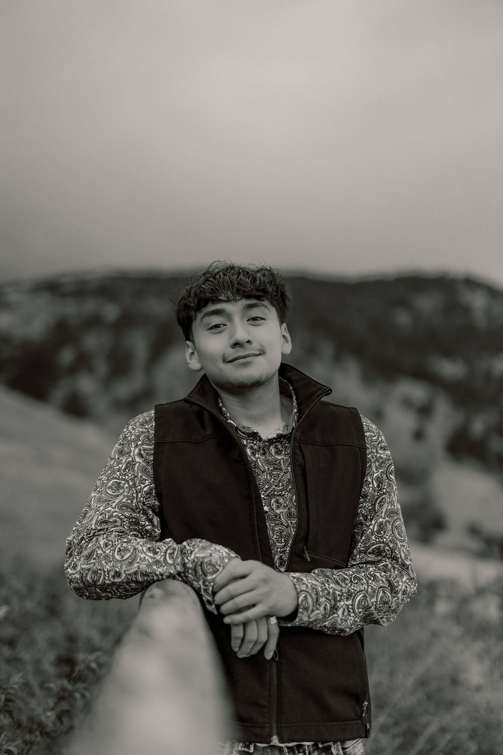 Black and white portrait of a young man leaning on a railing outdoors, wearing a patterned shirt and vest.