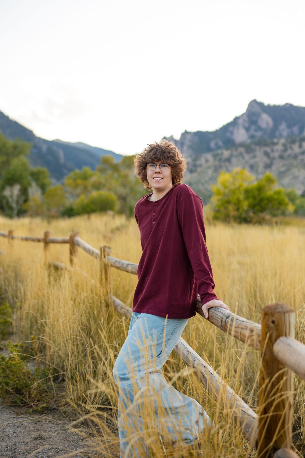 Person in burgundy sweater and light pants dances playfully in tall grass with mountains in background.