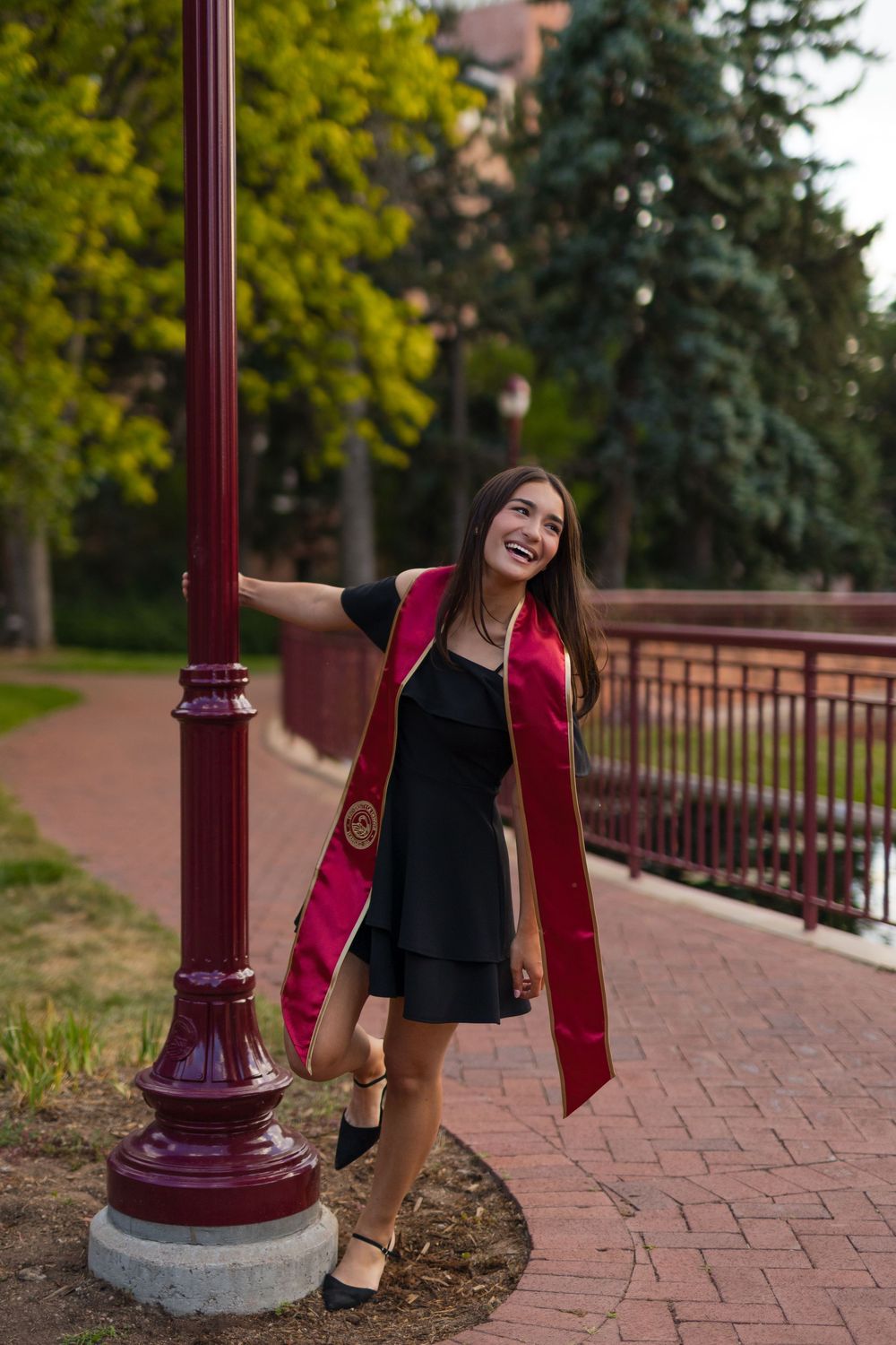 Smiling graduate in black dress and maroon sash poses by a lamppost on a brick campus walkway.