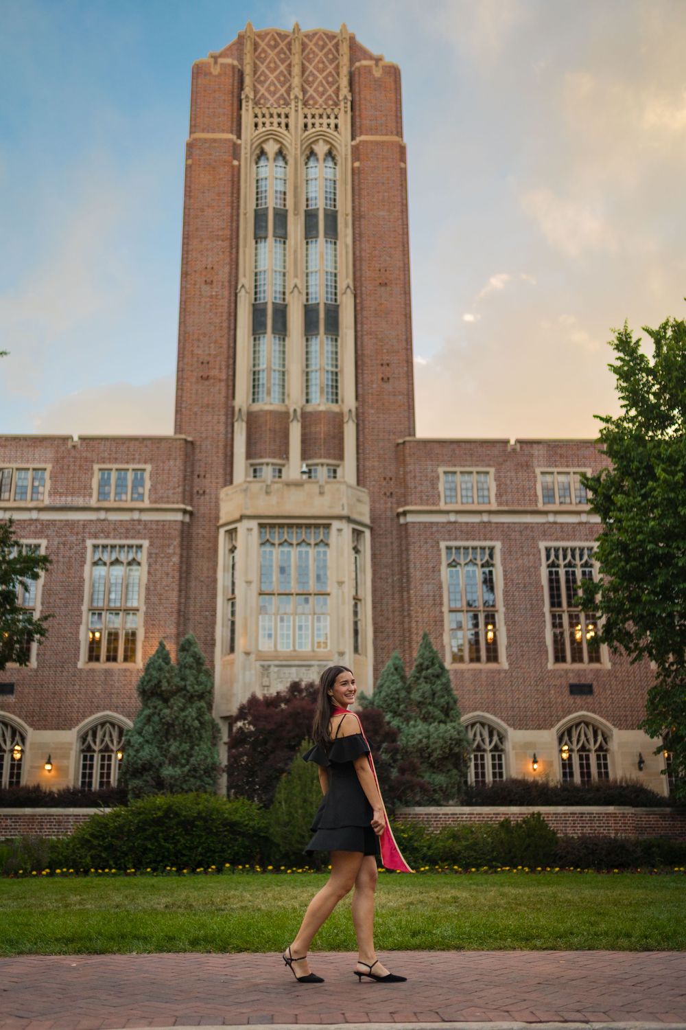 Woman in black dress walks in front of a grand Gothic brick university building tower at dusk.